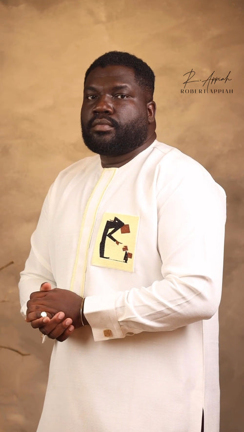Man in a white traditional outfit with a yellow embroidered design against a beige background